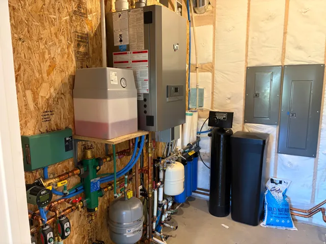 Utility area featuring a tankless water heater, comprehensive water filtration system, exposed PEX and copper plumbing, electrical panels, and unfinished plywood and insulated walls