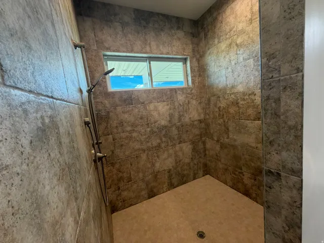 Tiled shower stall featuring textured wall tiles, a fixed showerhead with an attached handheld sprayer, and a small horizontal window