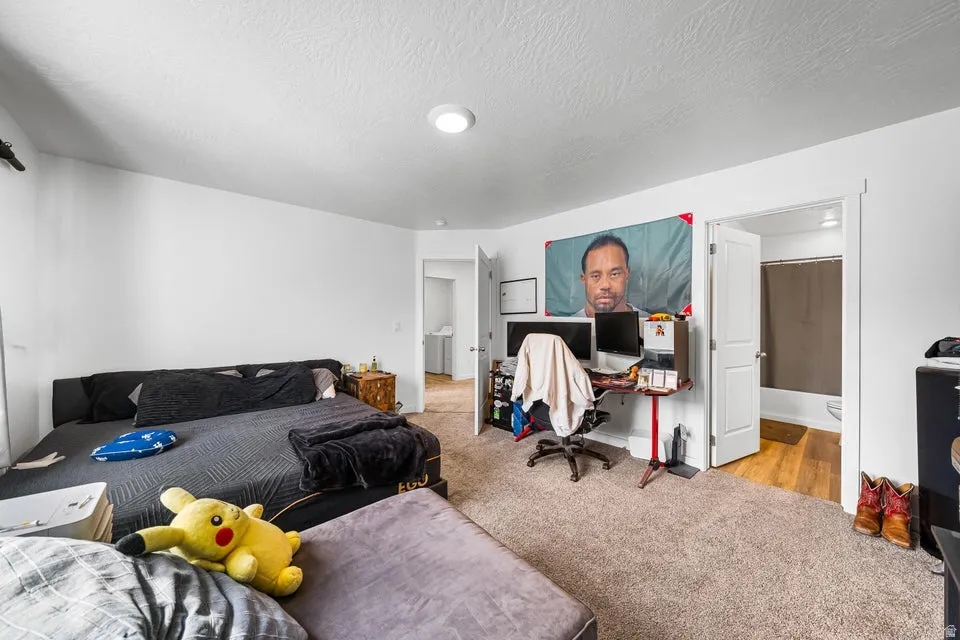 Spacious room with white walls, carpet flooring, a ceiling-mounted light fixture, and a doorway opening to a bathroom with wood-finish flooring and a tub/shower combination