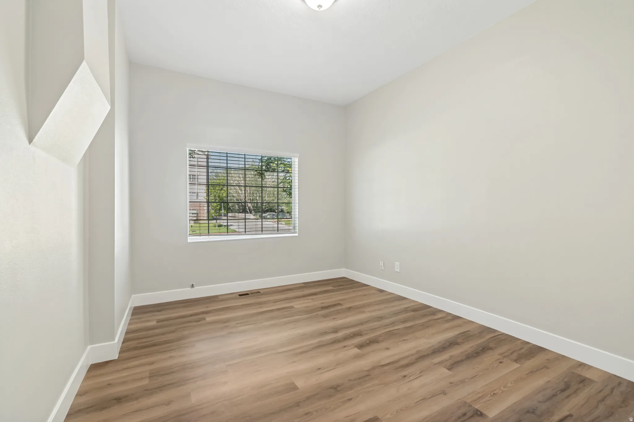Bright and spacious bedroom showcasing pristine white walls, abundant natural light from a large gridded window, and warm wood-finish flooring throughout