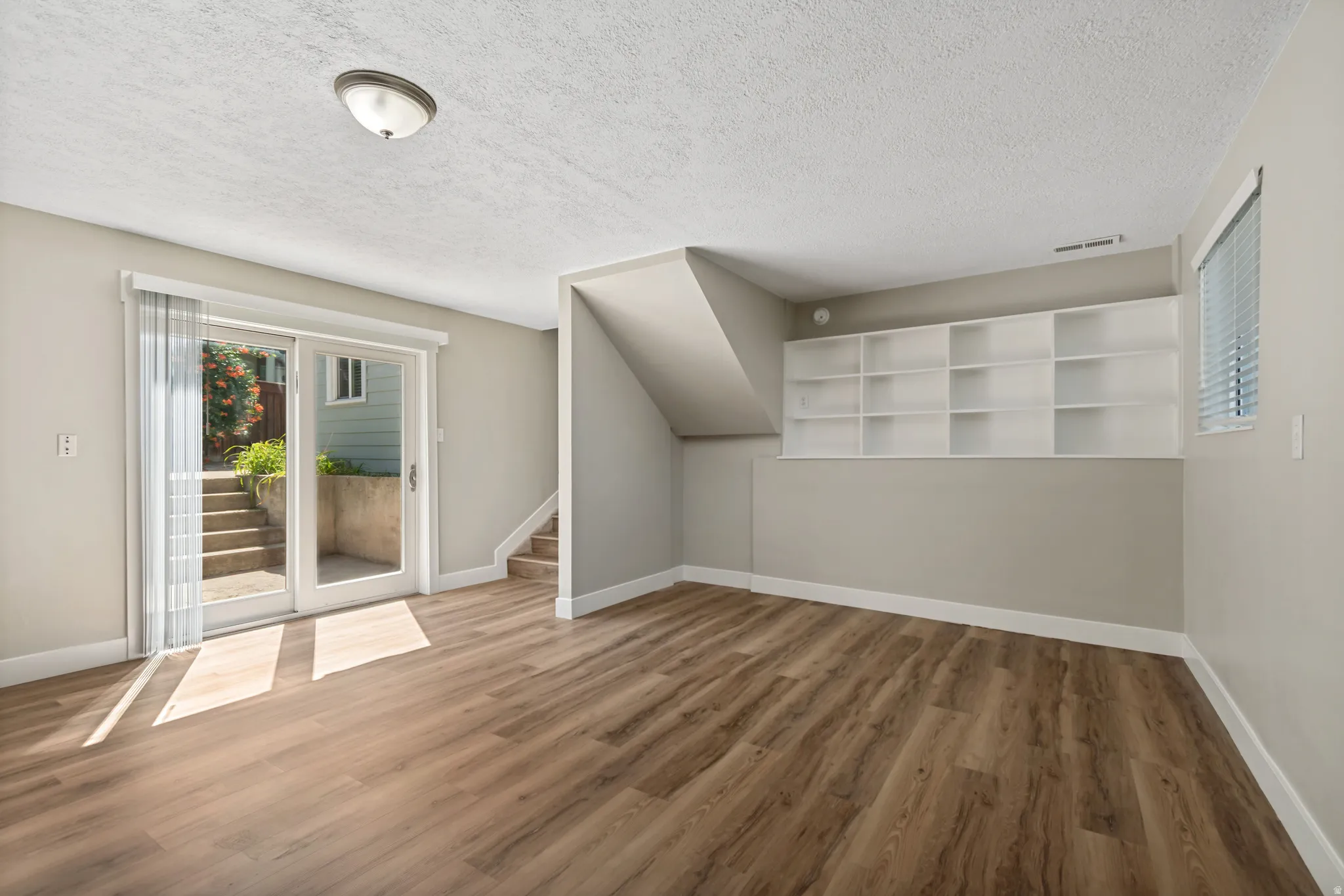 Spacious interior room featuring wood-finish flooring, built-in shelving, a large window with blinds, a ceiling-mounted light fixture, and glass sliding doors providing exterior access