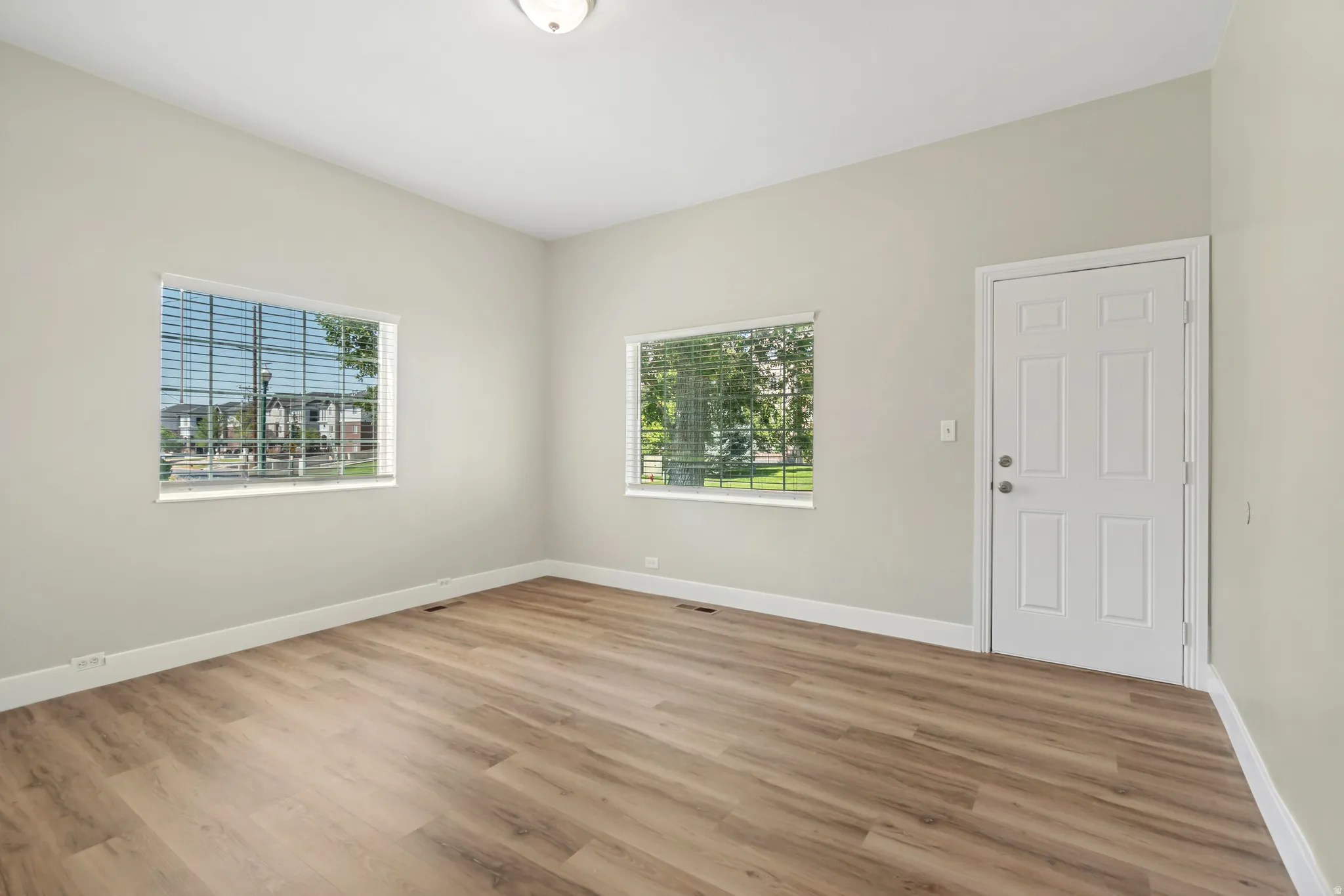 Bright and spacious room featuring gleaming wood-finish flooring, abundant natural light from two large windows, crisp white six-panel entry door, and fresh neutral wall finishes throughout