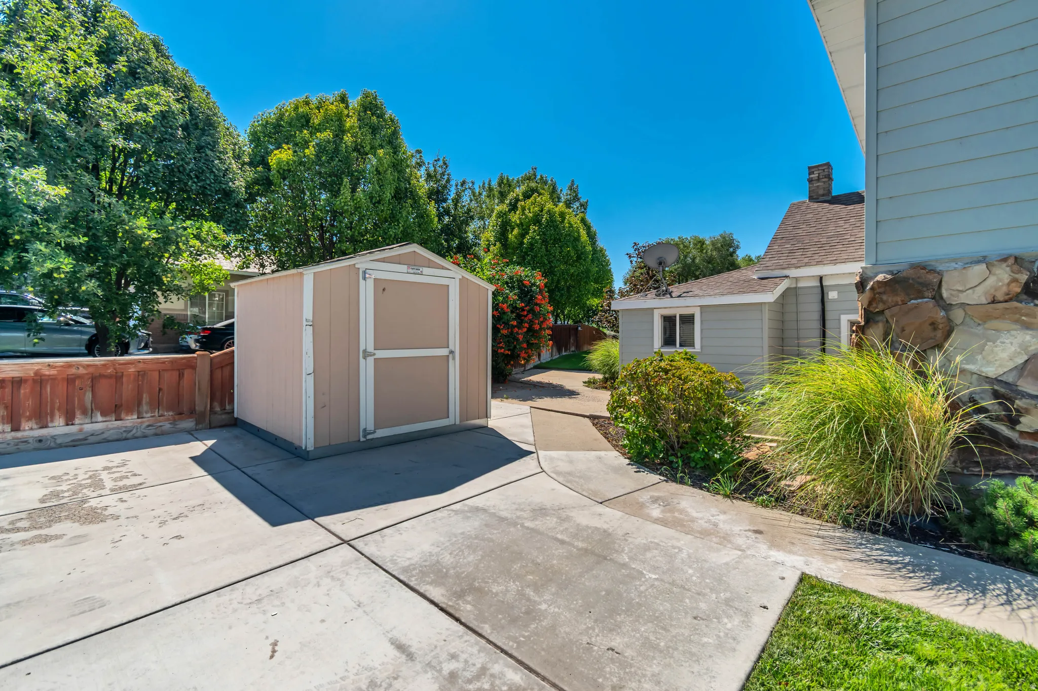 Spacious backyard featuring a versatile storage shed with dual-tone exterior and white trim, mature landscaping with ornamental grasses and established trees, concrete patio area, and adjacent single-story residence with siding exterior