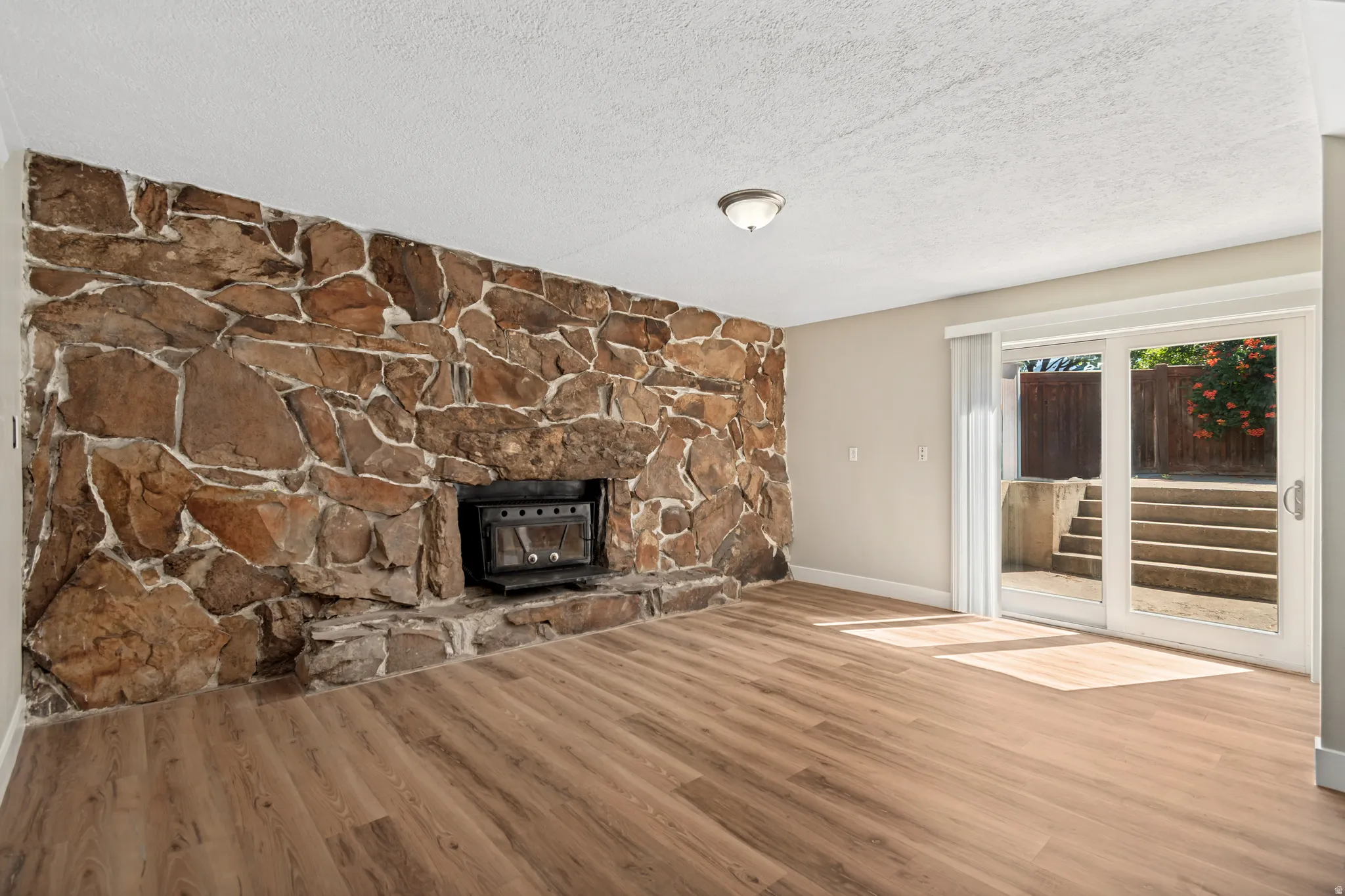 Spacious living area showcasing warm wood-finish flooring, a striking stone fireplace with integrated hearth, and abundant natural light from sliding glass doors opening to outdoor space