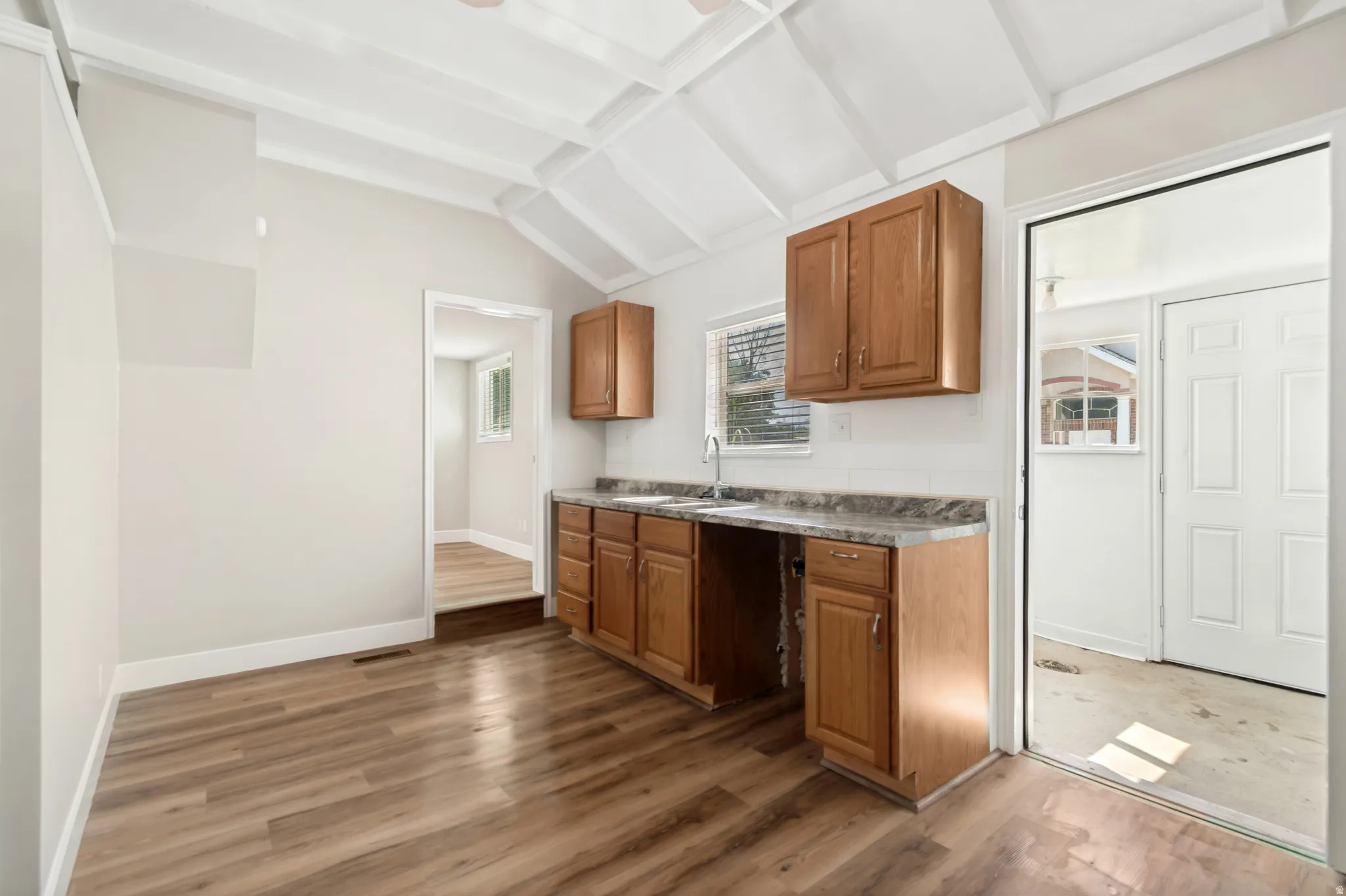 Kitchen area featuring vaulted ceilings, wood-finish flooring, a double basin sink, light wood cabinetry, and a countertop with a stone-look finish