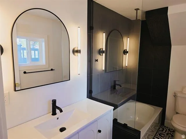 Bathroom featuring a single vanity with an undermount sink and matte black faucet, an arched mirror with sconce lighting, and a glass-enclosed tub/shower combination with dark tile surround