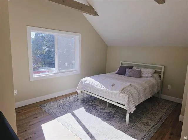 Bedroom featuring wood-finish flooring, a white trim window, sloped ceilings, and exposed wood beams