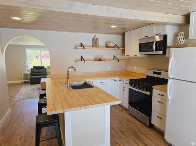 Modern kitchen featuring a large center island with wood-finish countertop and seating, white cabinetry with wood-tone accents, stainless steel appliances including a black range and microwave, bronze-finish faucet, wood-finish flooring throughout, and op