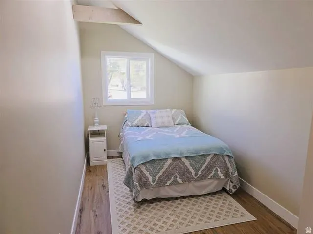 Attic room featuring an exposed ceiling beam, sloped ceiling, wood-finish flooring, a white-framed window, and neutral wall paint