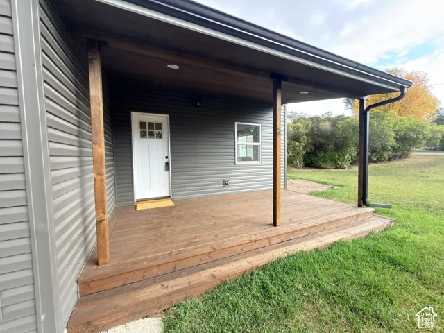 Covered exterior deck featuring natural wood-finish decking, stained wood support posts, dark horizontal siding, and a white six-panel door with inset glass