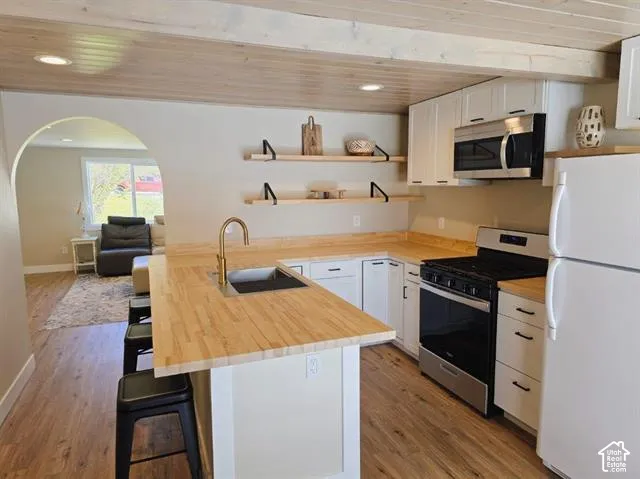 Modern kitchen featuring a large center island with wood-finish countertop and seating, white cabinetry with wood-tone accents, stainless steel appliances including a black range and microwave, bronze-finish faucet, wood-finish flooring throughout, and op