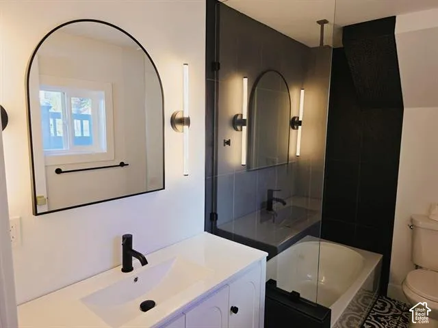 Bathroom featuring a single vanity with an undermount sink and matte black faucet, an arched mirror with sconce lighting, and a glass-enclosed tub/shower combination with dark tile surround