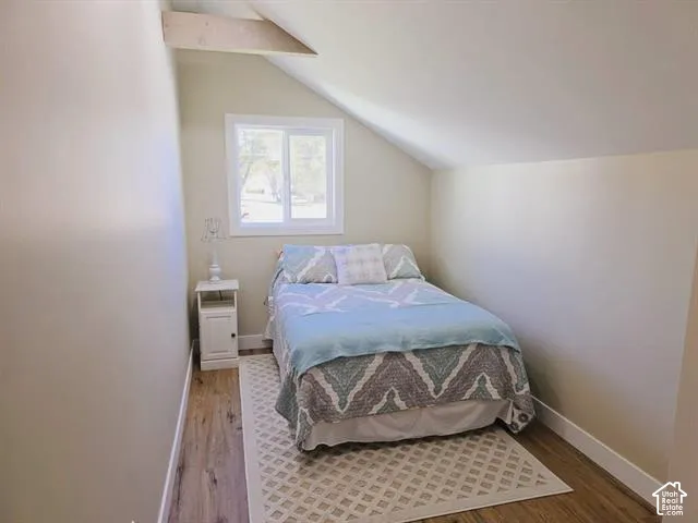 Attic room featuring an exposed ceiling beam, sloped ceiling, wood-finish flooring, a white-framed window, and neutral wall paint