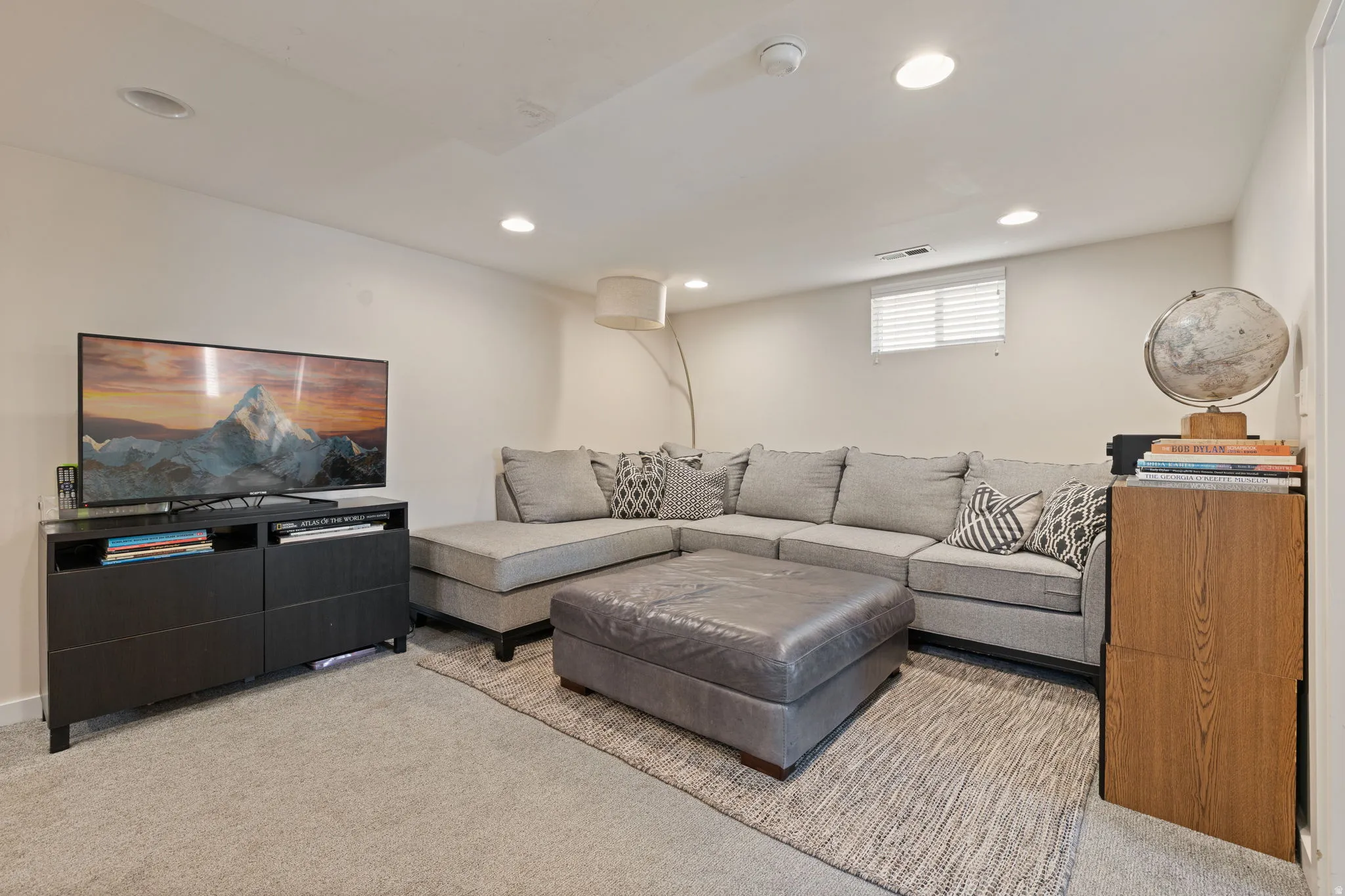 Finished basement recreation room featuring a sectional seating area, leather ottoman, recessed lighting throughout, and neutral carpet flooring