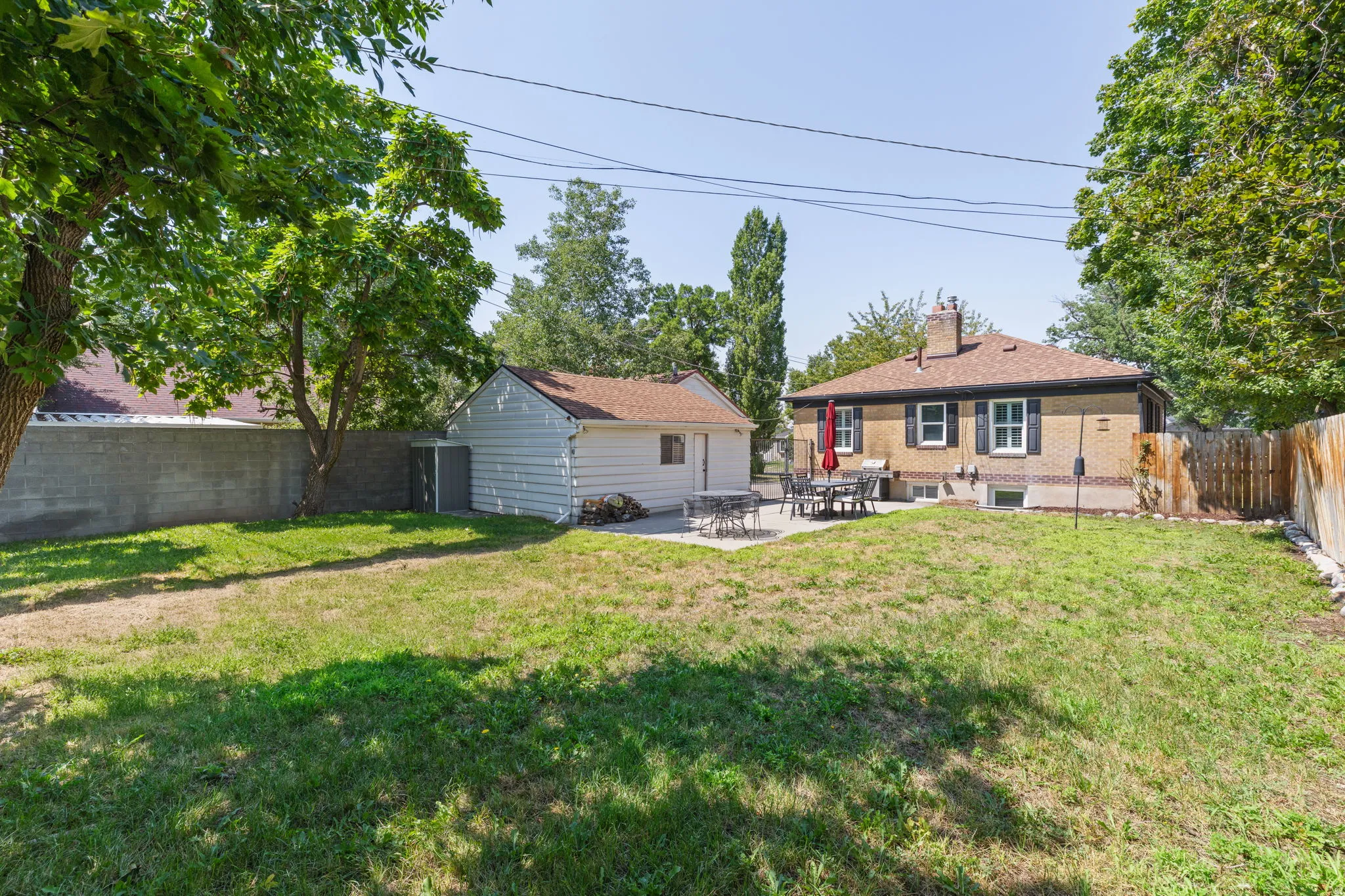 Spacious backyard featuring a mature tree-lined landscape with expansive lawn, detached garage structure, and single-story residence with yellow exterior siding and covered porch area