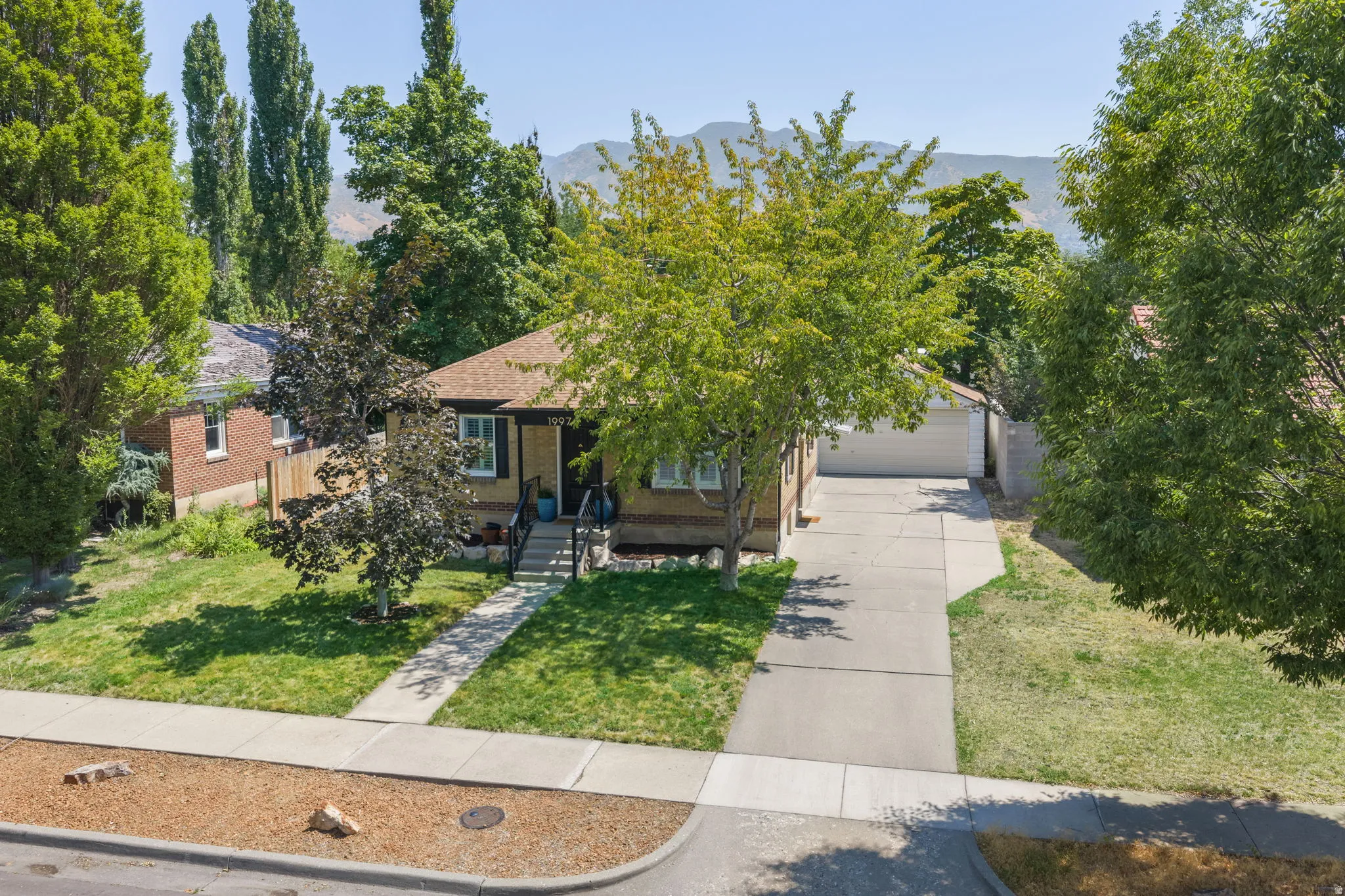 Charming brick residence showcasing a manicured front lawn, mature landscaping with poplar and shade trees, concrete driveway with attached garage, and a welcoming entry with covered porch