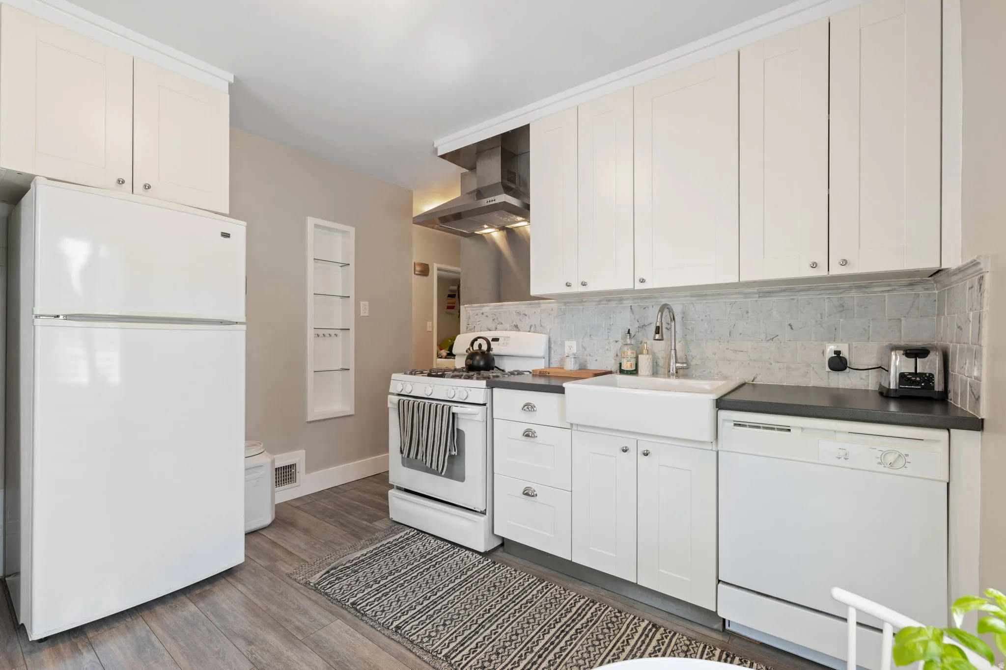 Kitchen featuring white upper and lower cabinetry, a white farmhouse sink, a tile backsplash, dark countertops, and wood-finish flooring
