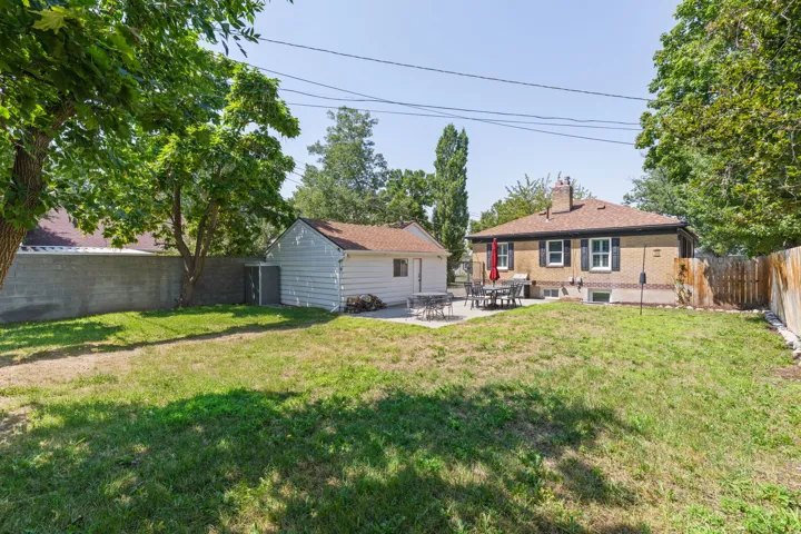 Spacious backyard featuring a mature tree-lined landscape with expansive lawn, detached garage structure, and single-story residence with yellow exterior siding and covered porch area