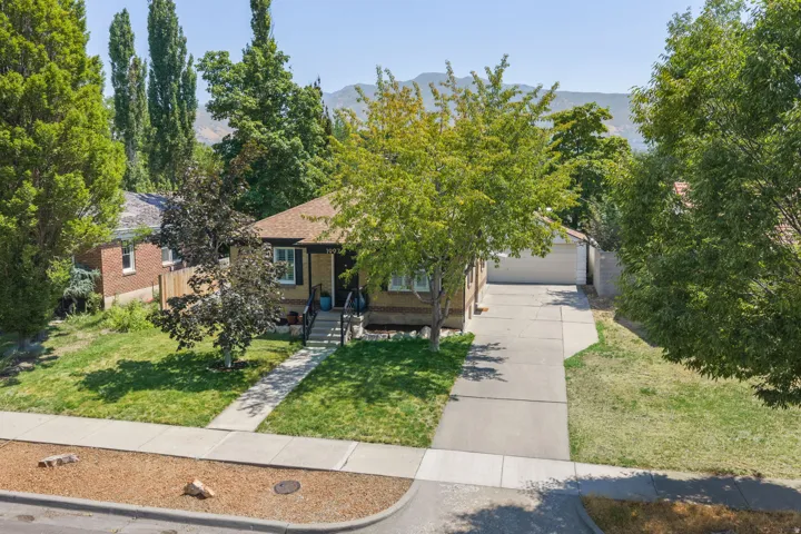 Charming brick residence showcasing a manicured front lawn, mature landscaping with poplar and shade trees, concrete driveway with attached garage, and a welcoming entry with covered porch