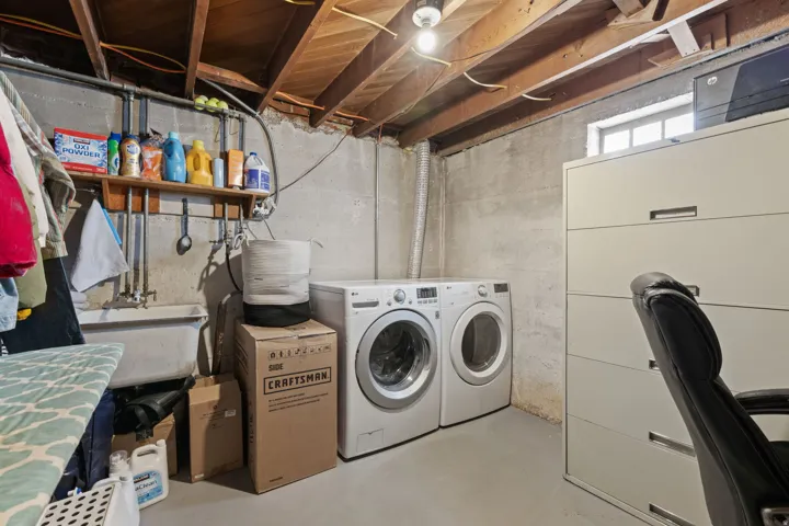 Dedicated laundry room featuring dual front-load washers and dryers, ample cabinetry for storage, exposed wooden beam ceiling, and concrete flooring with utility shelving for household supplies and equipment organization
