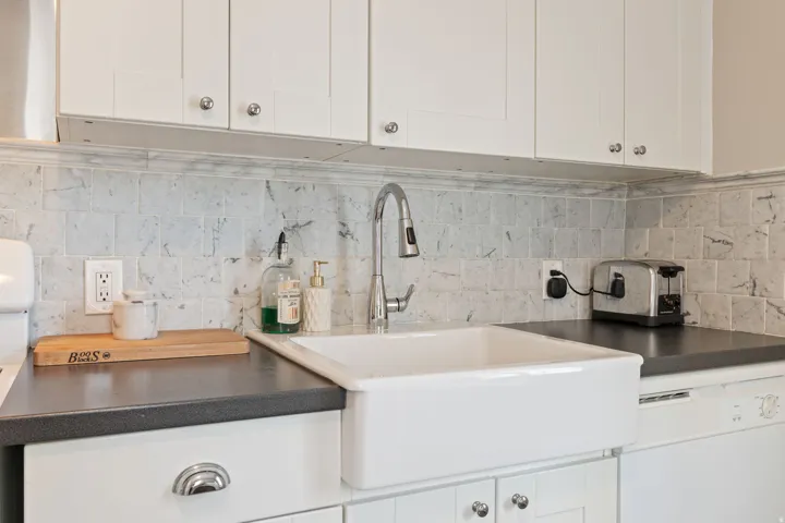 Kitchen featuring a white farmhouse sink with a gooseneck faucet, light-colored square tile backsplash, dark countertops, and white upper and lower cabinetry with chrome hardware