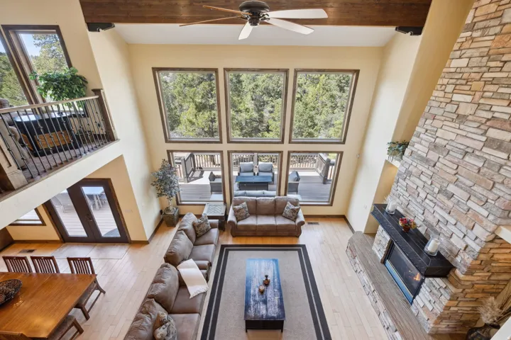 Soaring two-story great room showcases floor-to-ceiling multi-pane windows framing tree-lined scenery, hardwood flooring, a ceiling fan with exposed wood beams, a gas fireplace with stacked stone surround, and built-in shelving