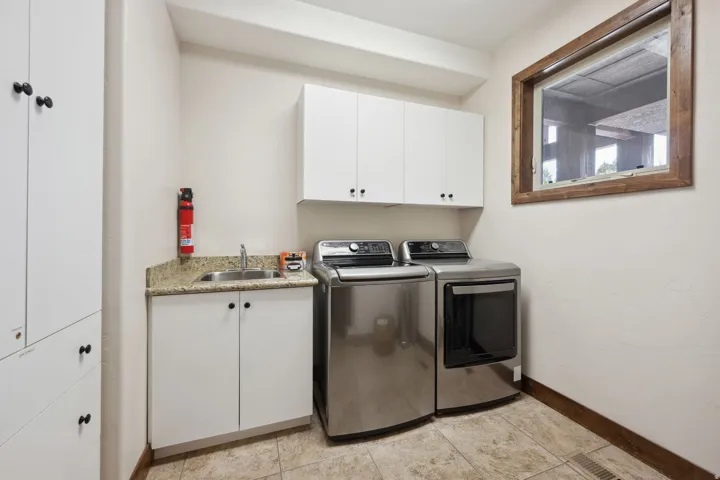 Dedicated laundry room featuring stainless steel front-load washer and dryer, white cabinetry with black hardware, granite-look countertop with undermount sink, and tile flooring