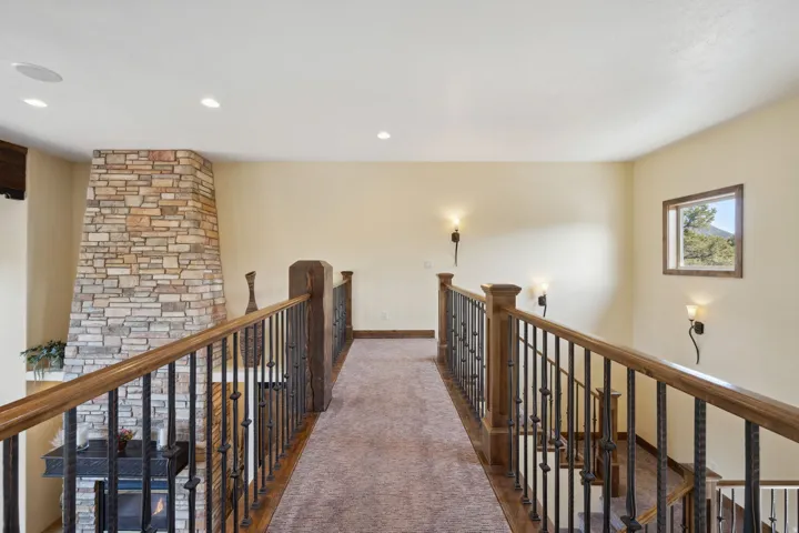 Carpeted hallway overlooking a stone-clad fireplace, featuring wood and wrought iron railings, recessed ceiling lighting, and wall sconces