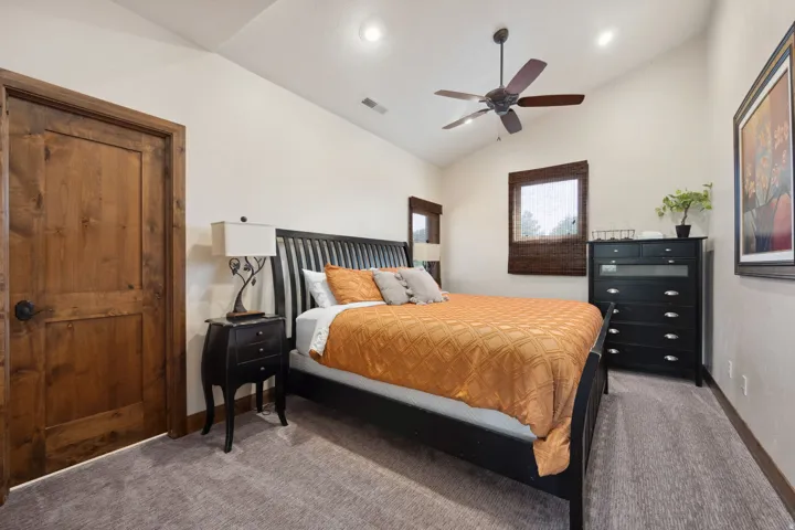 Spacious primary bedroom featuring a black metal bed frame with spindle headboard, warm wood-finish entry door, ceiling fan with dark blades, recessed lighting, and soft carpet flooring