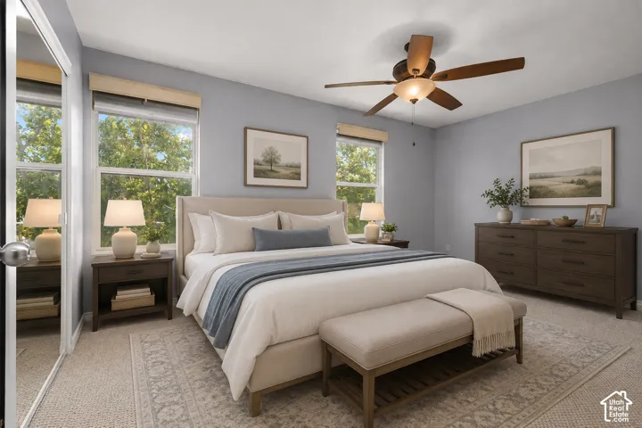Serene primary bedroom showcasing soft blue-gray walls, large multi-pane windows flooding the space with natural light, ceiling fan with integrated lighting, wood-finish nightstands with attached lamps, and a coordinating upholstered bench at the foot of