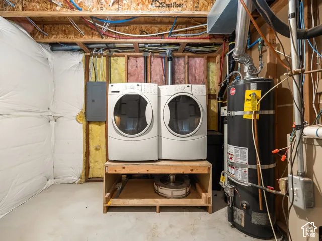 Dedicated laundry area featuring a white front-loading washer and dryer set, a hot water heater, exposed utility lines, and an electrical service panel
