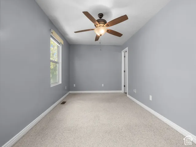 Bright and spacious bedroom featuring neutral gray walls, plush carpeting throughout, a ceiling fan with integrated lighting fixture, and natural light from a large window
