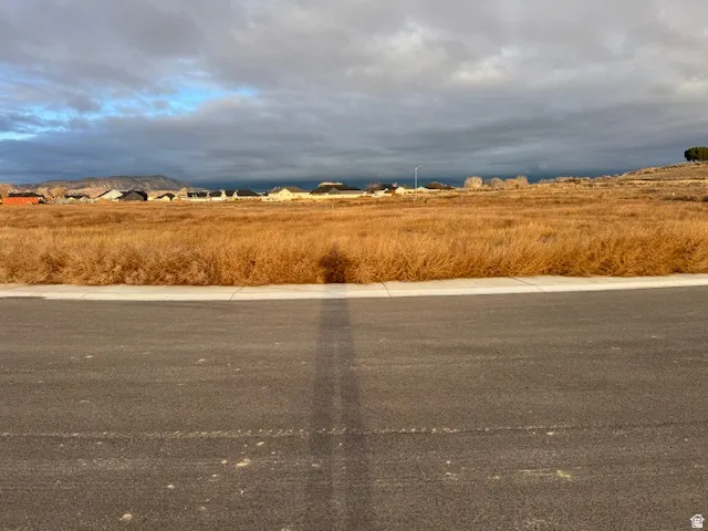 Expansive vacant land parcel featuring a paved street, concrete sidewalk, and distant residential structures