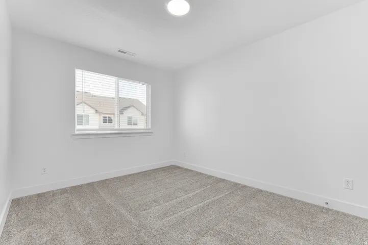 Bright secondary bedroom with expansive carpet flooring, large double-hung window with plantation shutters providing natural light, and fresh white wall finishes throughout