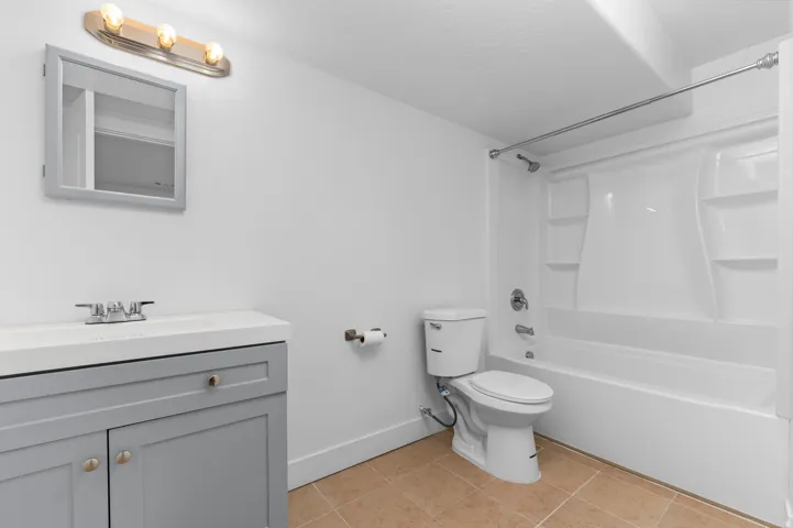 Contemporary bathroom featuring a light gray vanity with a white countertop, a mirrored medicine cabinet, a three-light vanity fixture, a white toilet, and a white tub with an integrated shower unit
