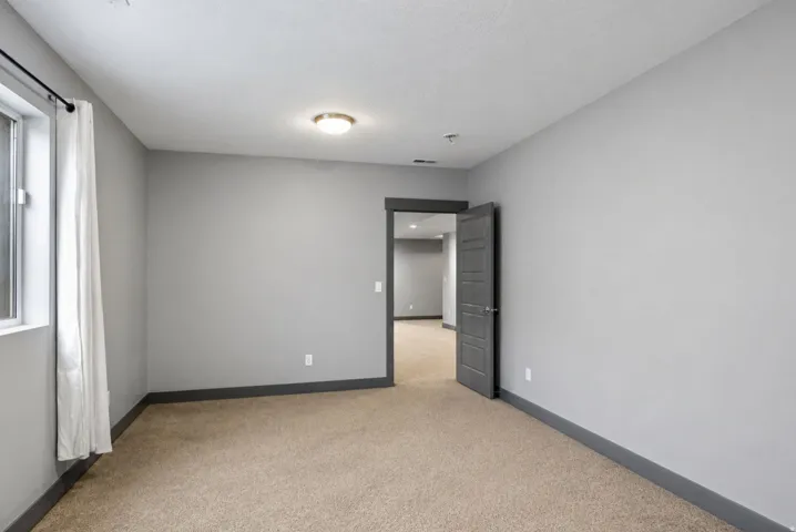 Neutral gray wall paint, brown carpet flooring, flush mount ceiling light fixture, white window trim, and a dark gray interior door