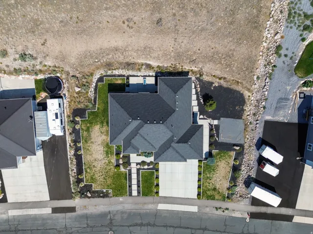 Aerial view showcasing a modern residential home featuring a distinctive multi-level roof design with dark asphalt shingles, integrated landscaping with manicured lawn sections, concrete hardscaping and pathways, and a compact footprint ideal for urban or