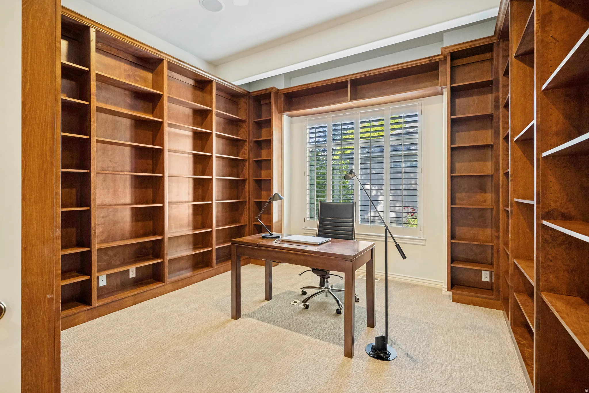 Office featuring extensive built-in wooden shelving, a large window with plantation shutters, and light-toned carpeting