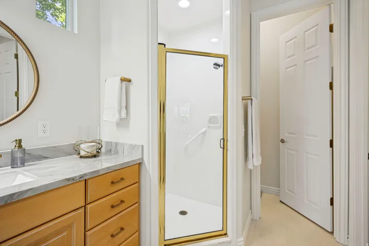 Bathroom featuring a glass-enclosed shower with gold-finish hardware, a double vanity with a quartz stone countertop, maple cabinetry, tastefully framed mirrors.