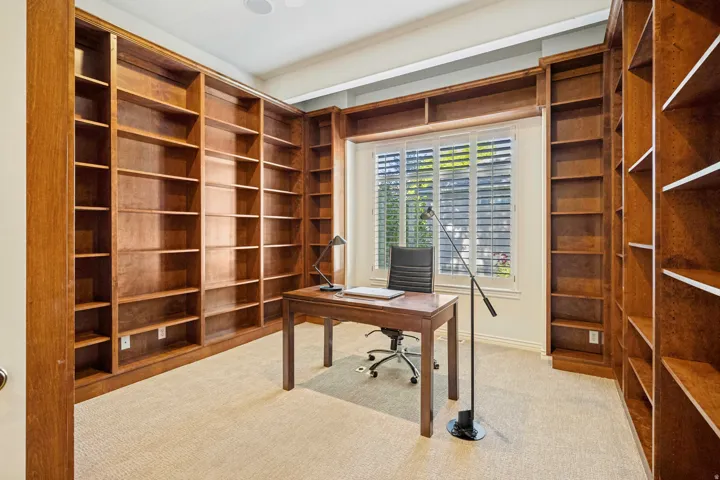 Office featuring extensive built-in wooden shelving, a large window with plantation shutters, and light-toned carpeting