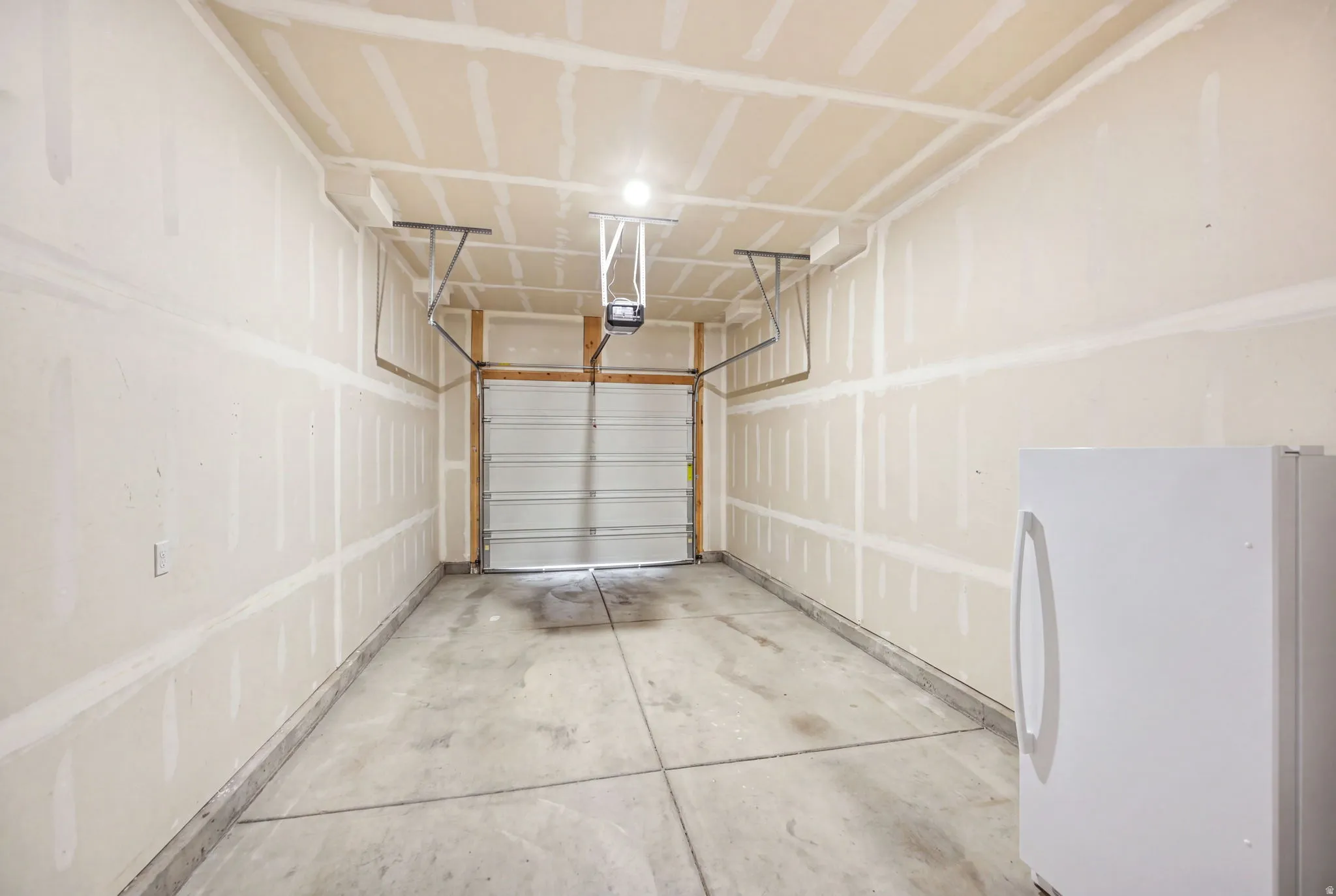 Spacious garage featuring concrete flooring, overhead garage door with automatic opener, wall-mounted basketball hoop, fluorescent ceiling lighting, and finished drywall construction throughout