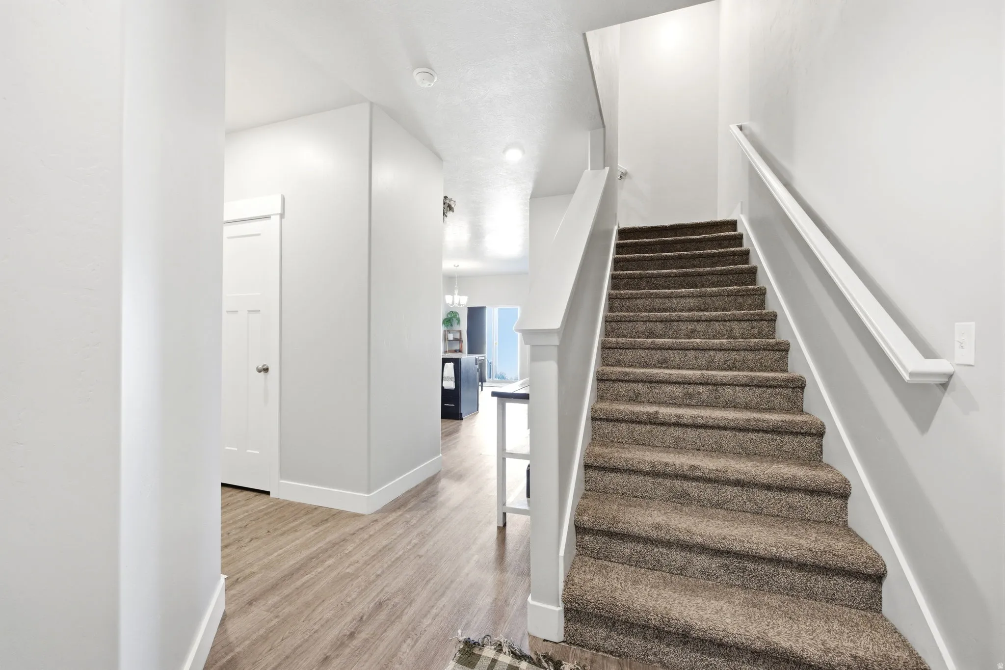 Bright hallway and staircase featuring wood-finish flooring, clean white walls, and a carpeted staircase with white railings and handrails