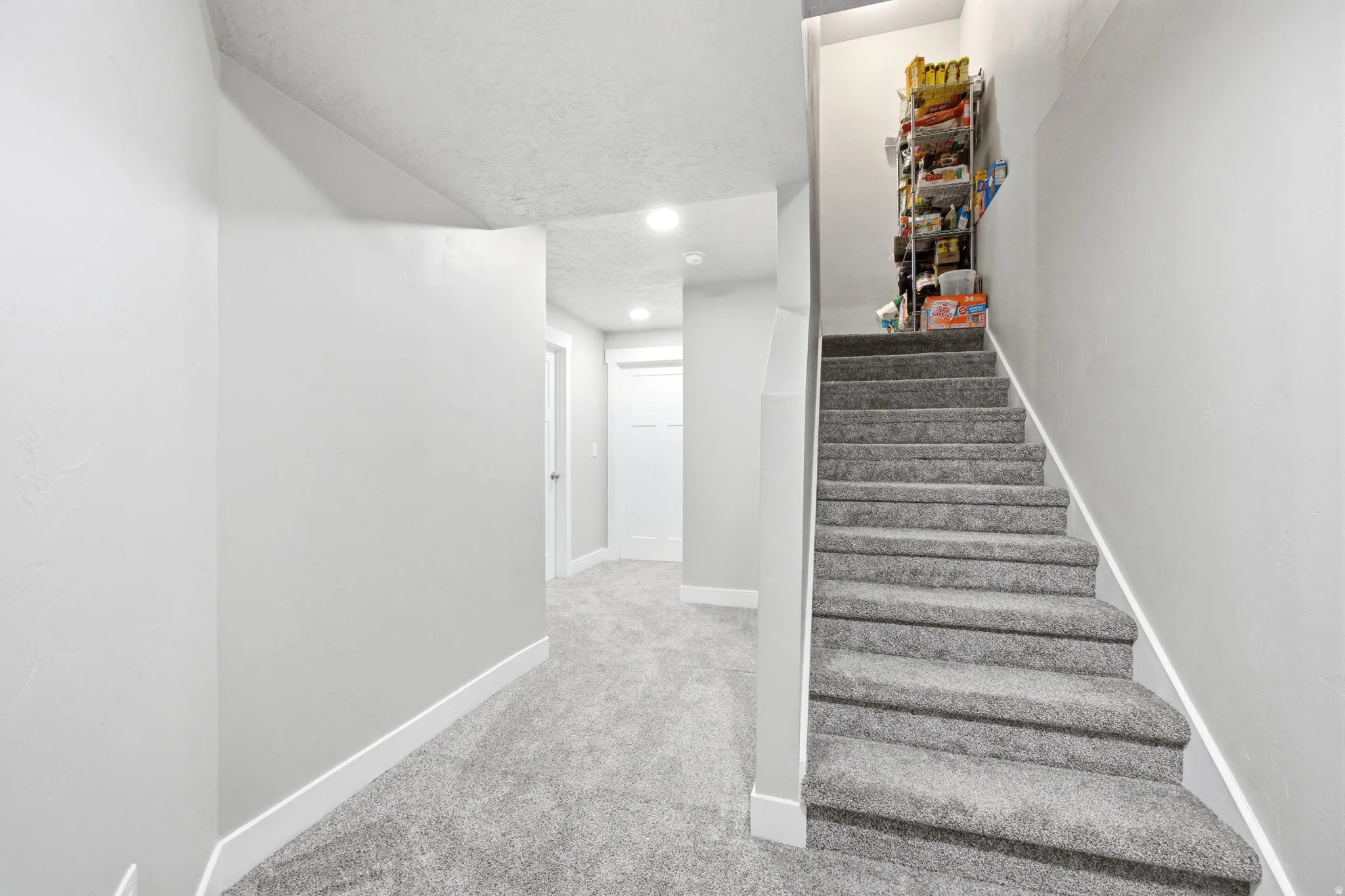 Bright hallway and staircase featuring neutral carpet flooring, recessed ceiling lighting, white painted walls, and a sturdy handrail