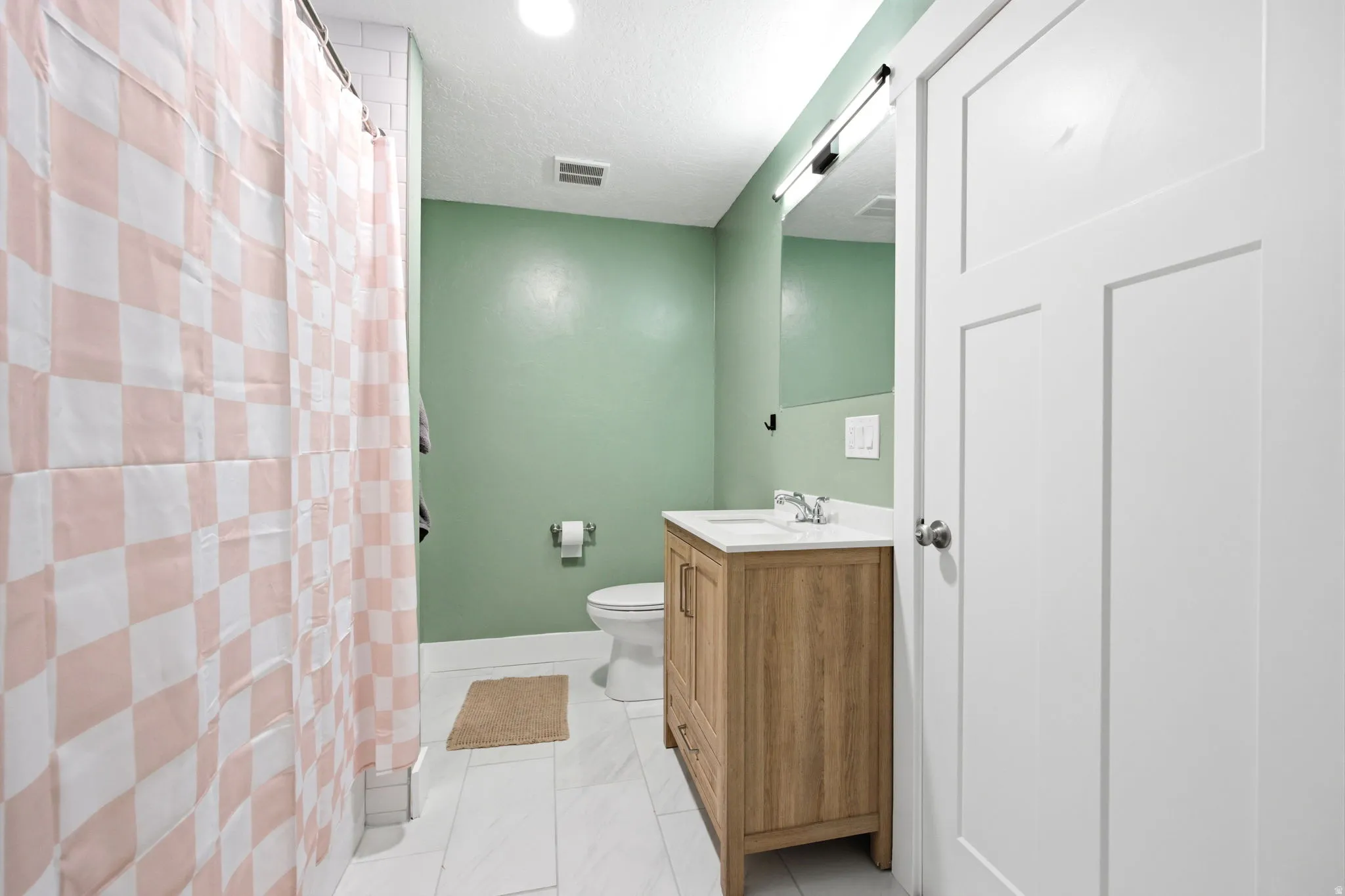 Modern primary bathroom featuring soft green accent walls, a wood-finish vanity with integrated sink and countertop, white subway tile flooring, and a pink and white checkered tile shower enclosure with glass door