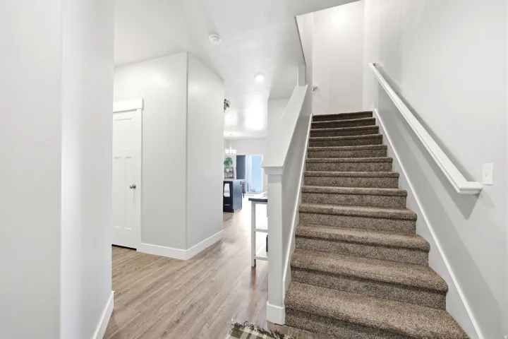 Bright hallway and staircase featuring wood-finish flooring, clean white walls, and a carpeted staircase with white railings and handrails
