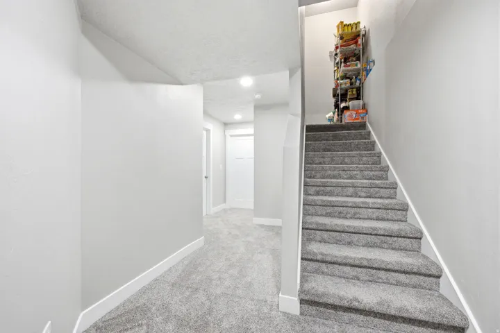 Bright hallway and staircase featuring neutral carpet flooring, recessed ceiling lighting, white painted walls, and a sturdy handrail