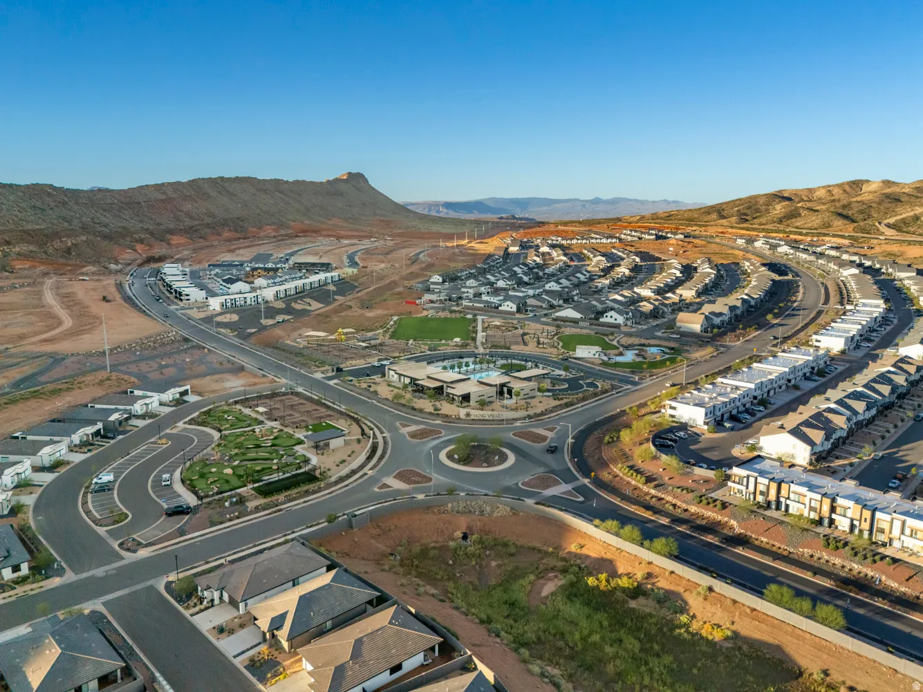 Expansive residential development featuring a central roundabout, numerous residential units, a community park with green space, a recreational pool area, and surrounding natural desert landscape