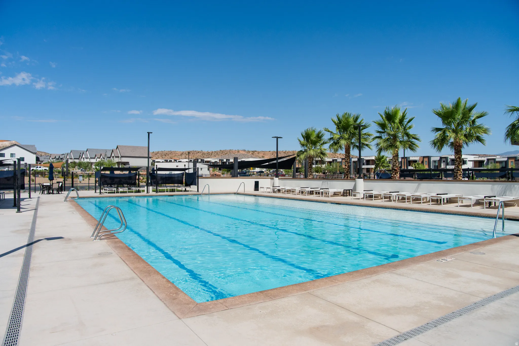 Community swimming pool with clear water, surrounded by concrete decking, palm trees, and modern residential structures