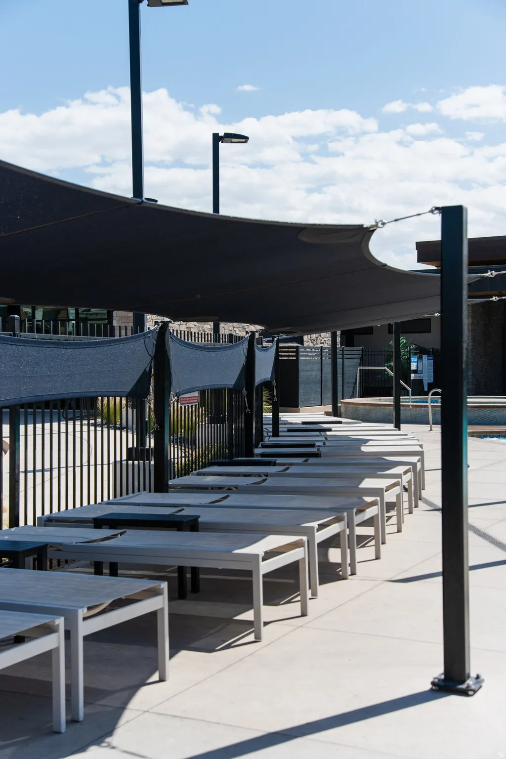 Poolside lounge area featuring a large shade sail, numerous lounge chairs, concrete decking, and modern light fixtures
