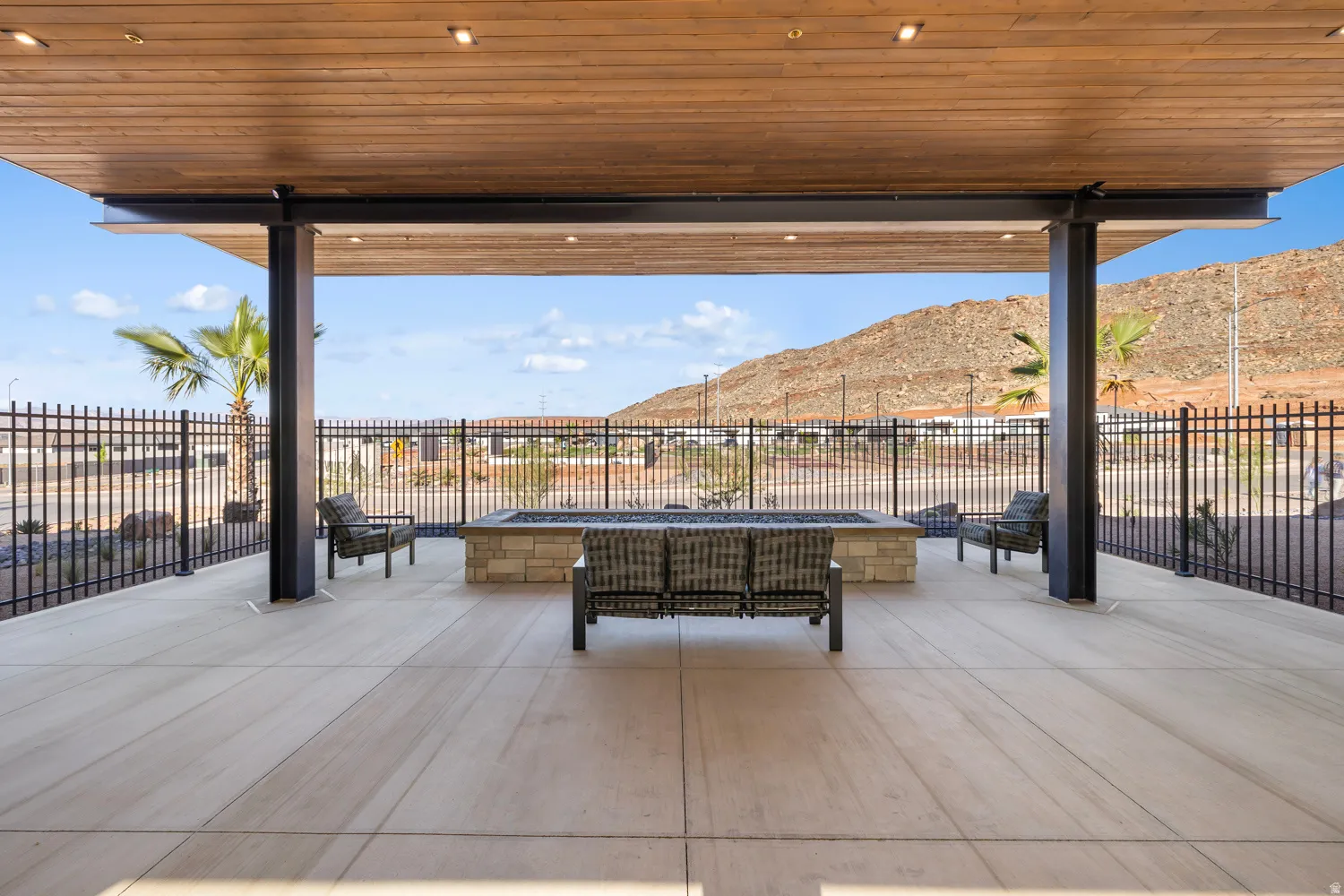 Expansive covered patio featuring a wood-finish ceiling with recessed lighting, large format concrete pavers, a built-in stone fire pit, and metal perimeter fencing